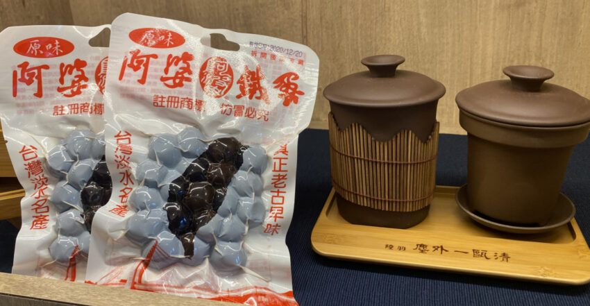 淡水名產｜#2 阿婆鐵蛋創始店｜鐵蛋的由來你知道嗎？故事是這樣的，女主角這位阿婆本身在經營一間麵店，某年雨季特別長，她滷了的滷蛋，因為天氣關係賣不出去，為避免浪費，老闆娘便一次又一次的回滷，結果魯蛋體積開始縮水、變得又小又黑。起初，有些客人對這個又小、又黑、又硬的滷蛋感到好奇，就買來吃看看，後來發現這個「黑金蛋」，咀嚼時帶著蛋香、口感極有嚼勁，在口耳相傳的力量下，鐵蛋便成為淡水超人氣伴手禮了！